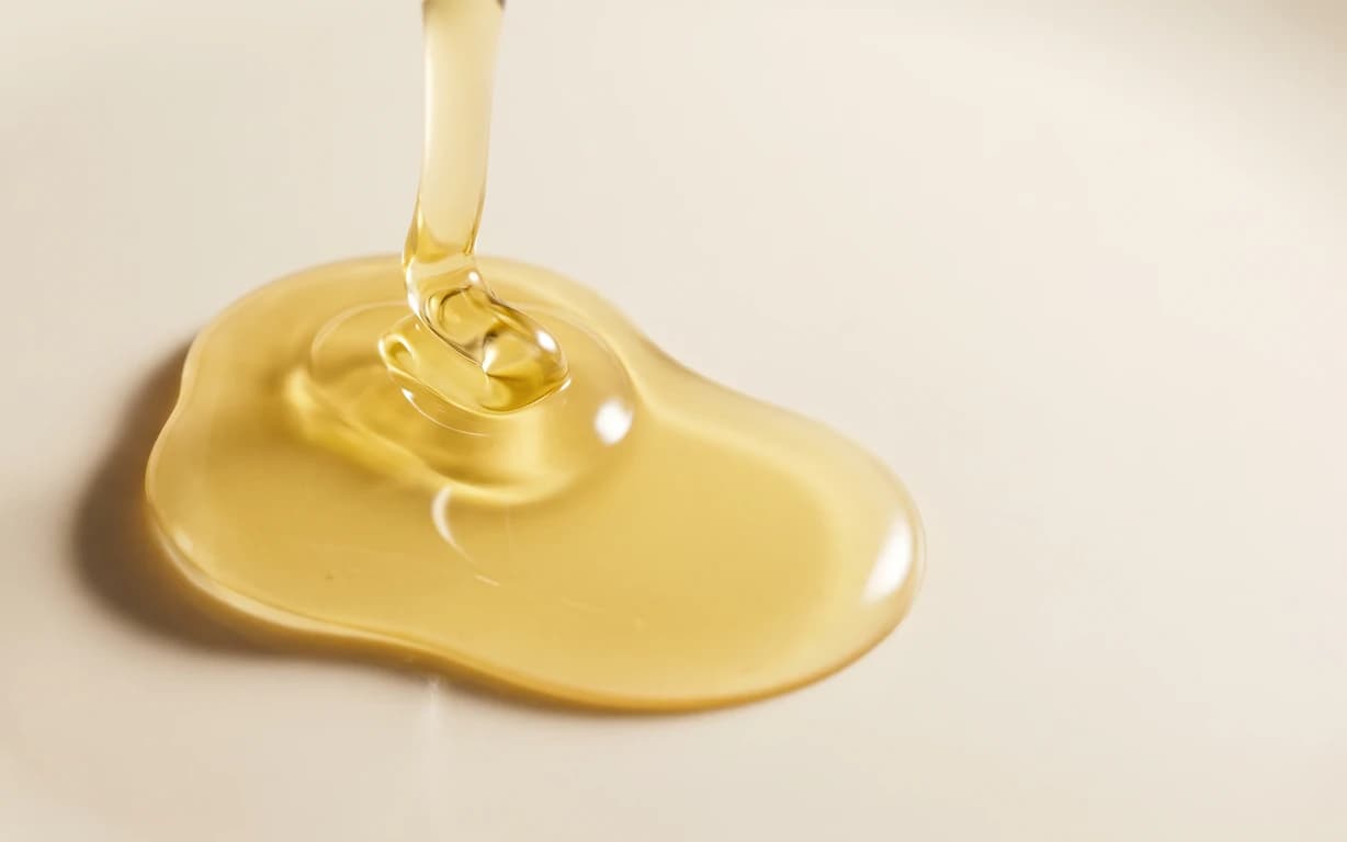 Golden oil pours into a glossy pool on a warm ivory surface, with soft highlights, a gentle shadow and ample negative space.
