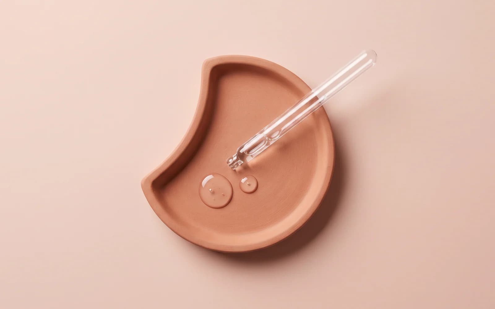 A clear glass pipette across a terracotta crescent tray beside two transparent droplets.