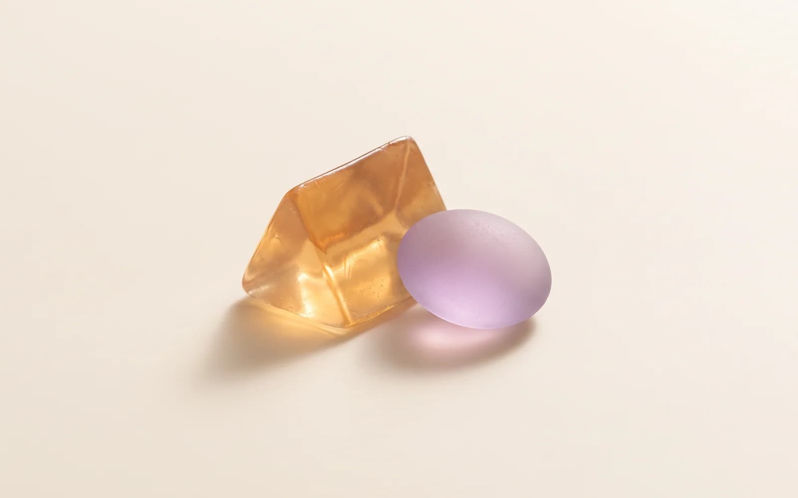An amber gel wedge beside a frosted lilac glass pebble on ivory.