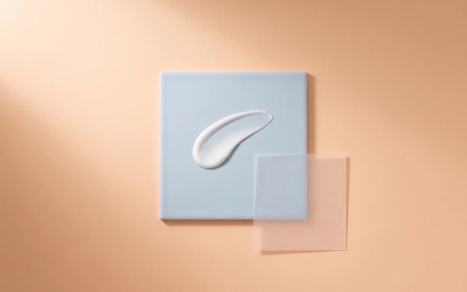 A thin white lotion stroke on a frosted blue tile beside a translucent film.