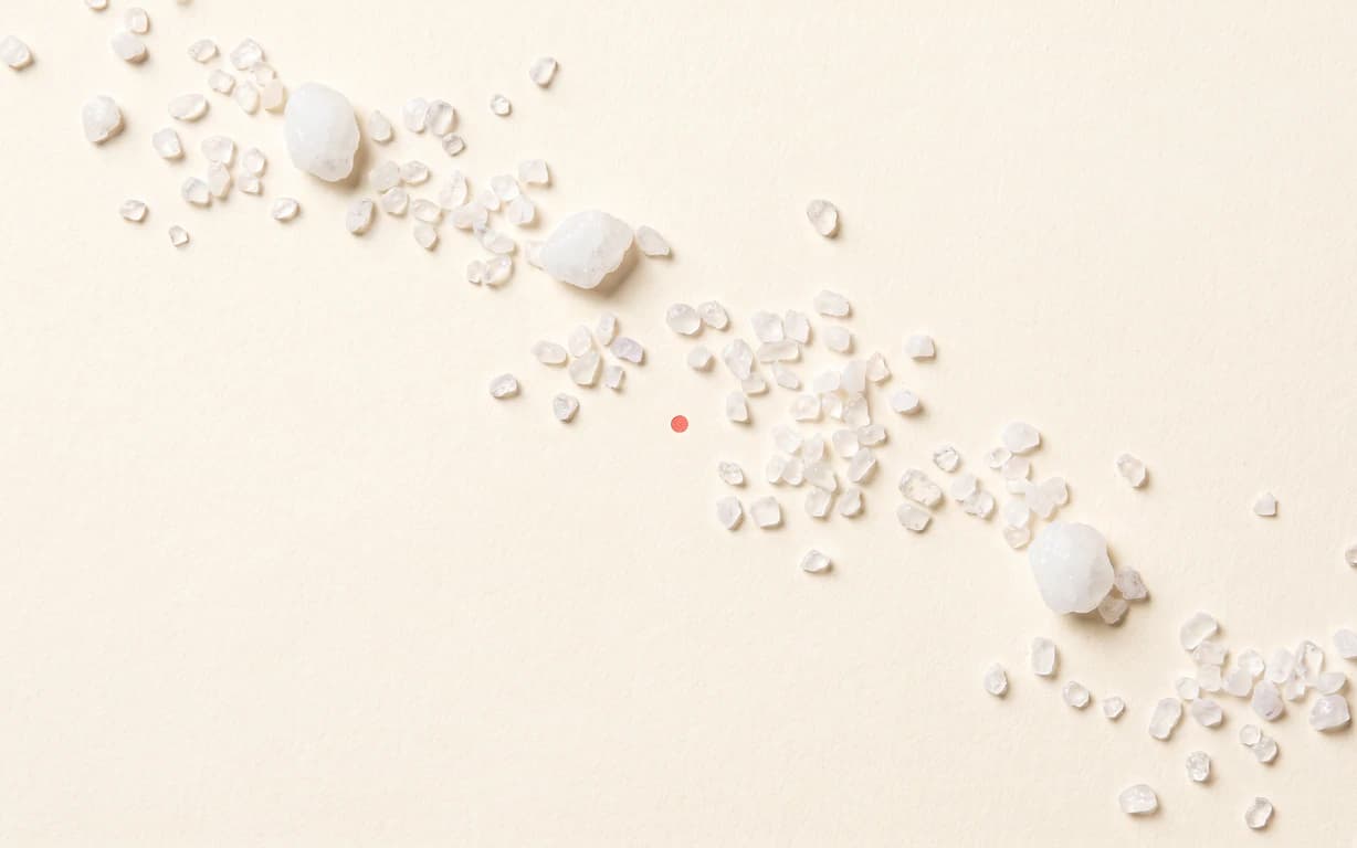 A loose diagonal trail of small milky xylitol crystals crosses a warm-ivory surface beside a tiny coral point.