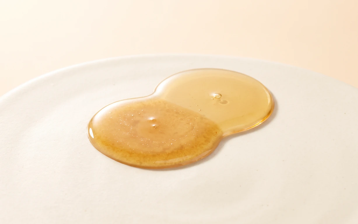 Two adjoining pools of translucent golden oil, one smooth and one lightly textured, resting on a softly lit ivory surface against a warm cream backdrop.