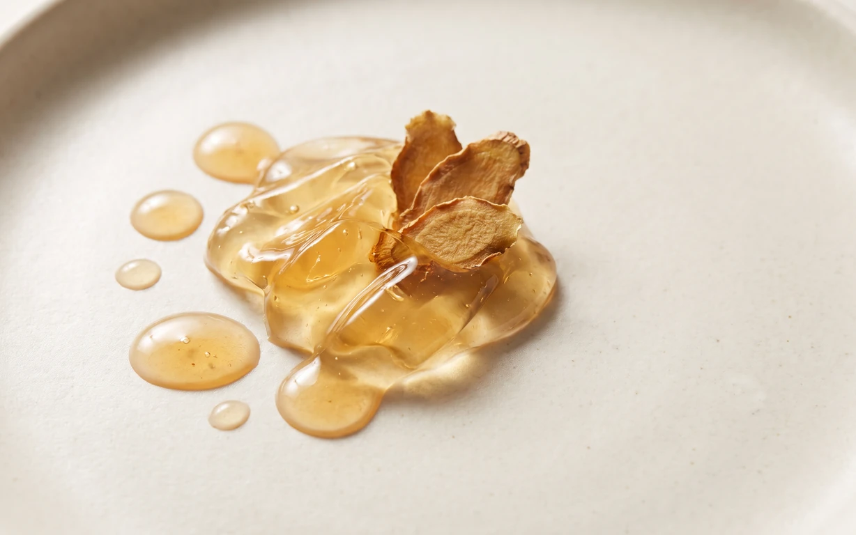 Amber translucent gel and droplets topped with dried ginger root slices on a softly lit ivory surface.