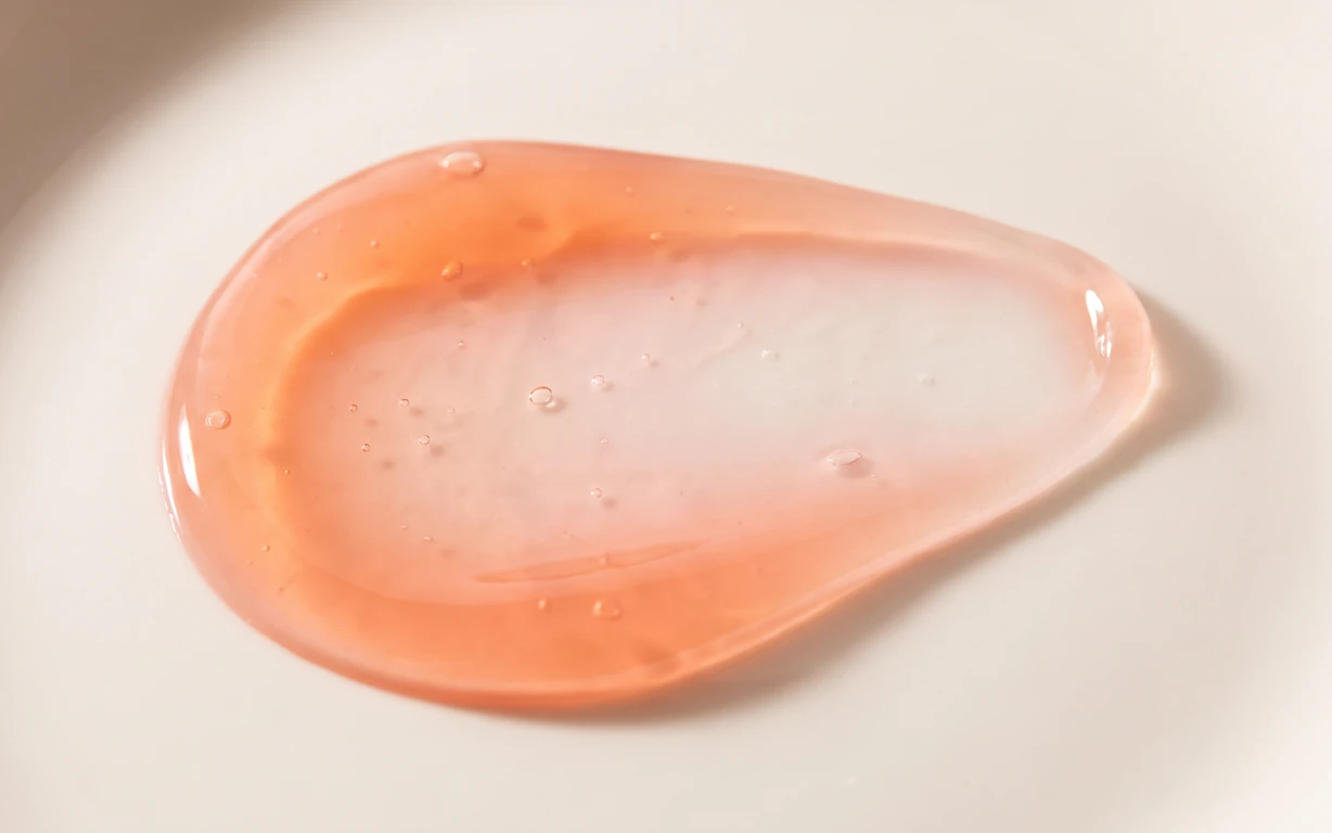 A translucent peach-colored abstract teardrop form with small droplets, softly lit on an ivory surface.
