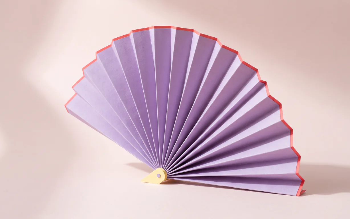 Pleated lavender paper fan with a coral-red edge on a pale blush surface.