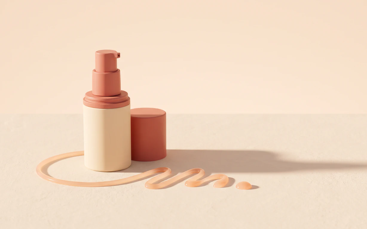 A label-free ivory and coral airless pump bottle stands beside its matching cap and a glossy peach serum ribbon on a warm ivory surface.
