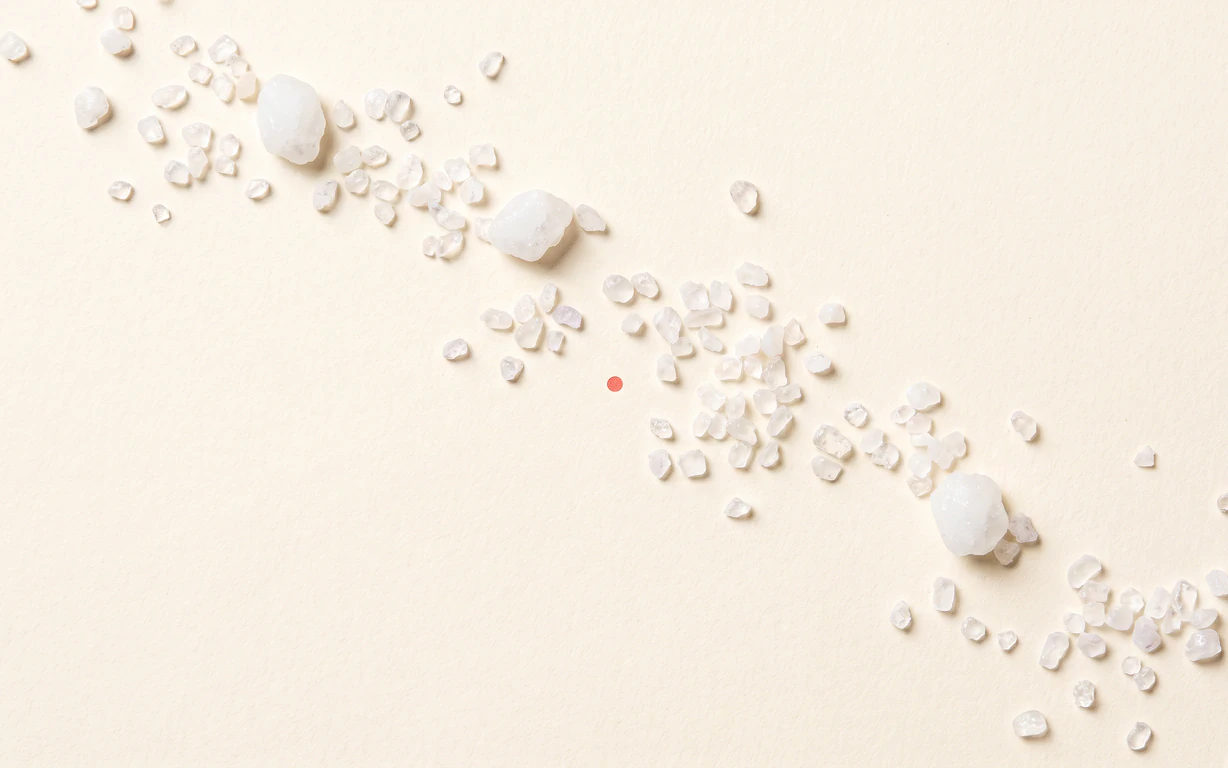 A loose diagonal trail of small milky xylitol crystals crosses a warm-ivory surface beside a tiny coral point.